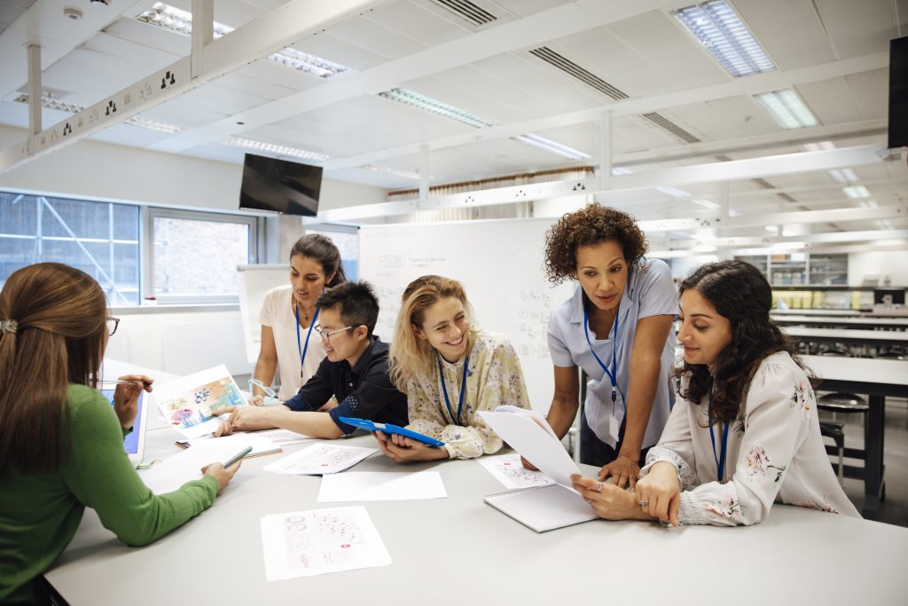  A group of students are sitting around a table in a classroom. They are working on a project together. The teacher is standing next to them, helping them with their work.