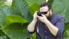 An image of a bearded man with sunglasses taking a picture with a camera. There are giant leaf's behind him.