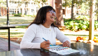 An image of a woman with dark hair and glasses painting a watercolor while looking up and laughing. 