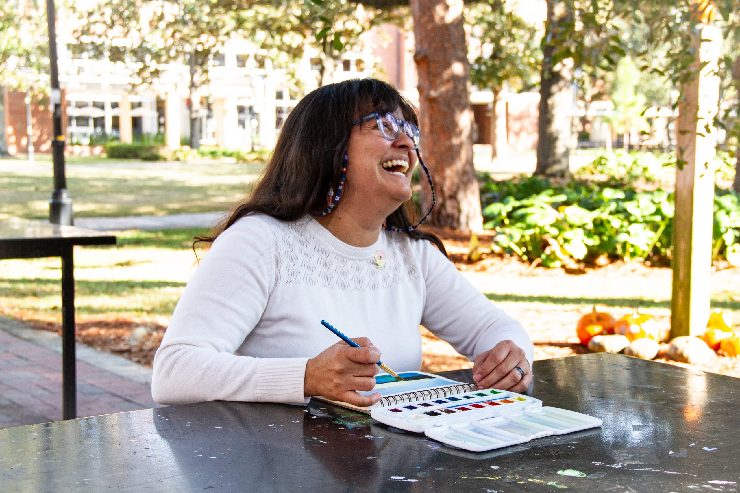 An image of a woman with dark hair and glasses painting a watercolor while looking up and laughing. 