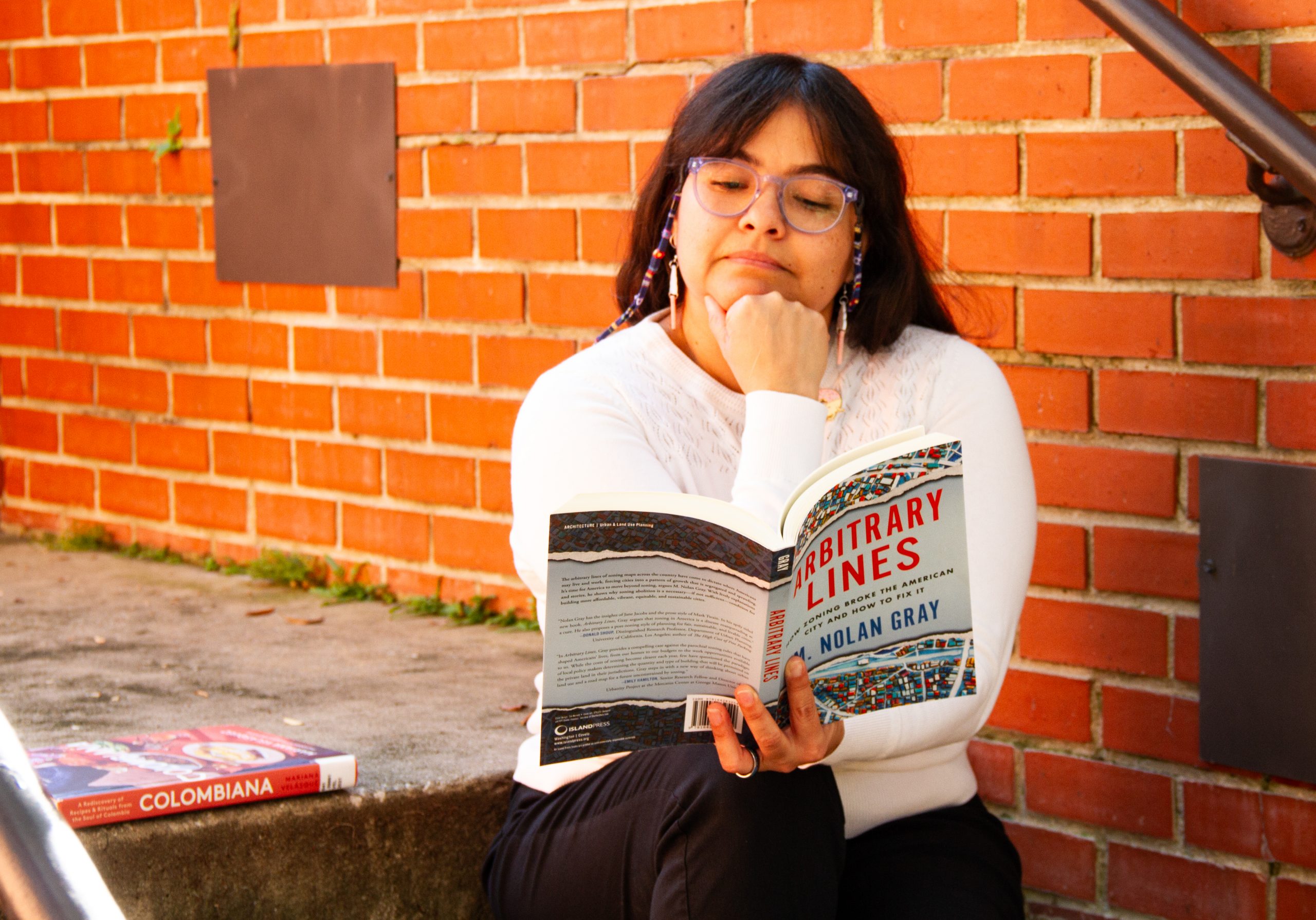 An image of a woman reading a book on the stairs with a brick wall behind her. 