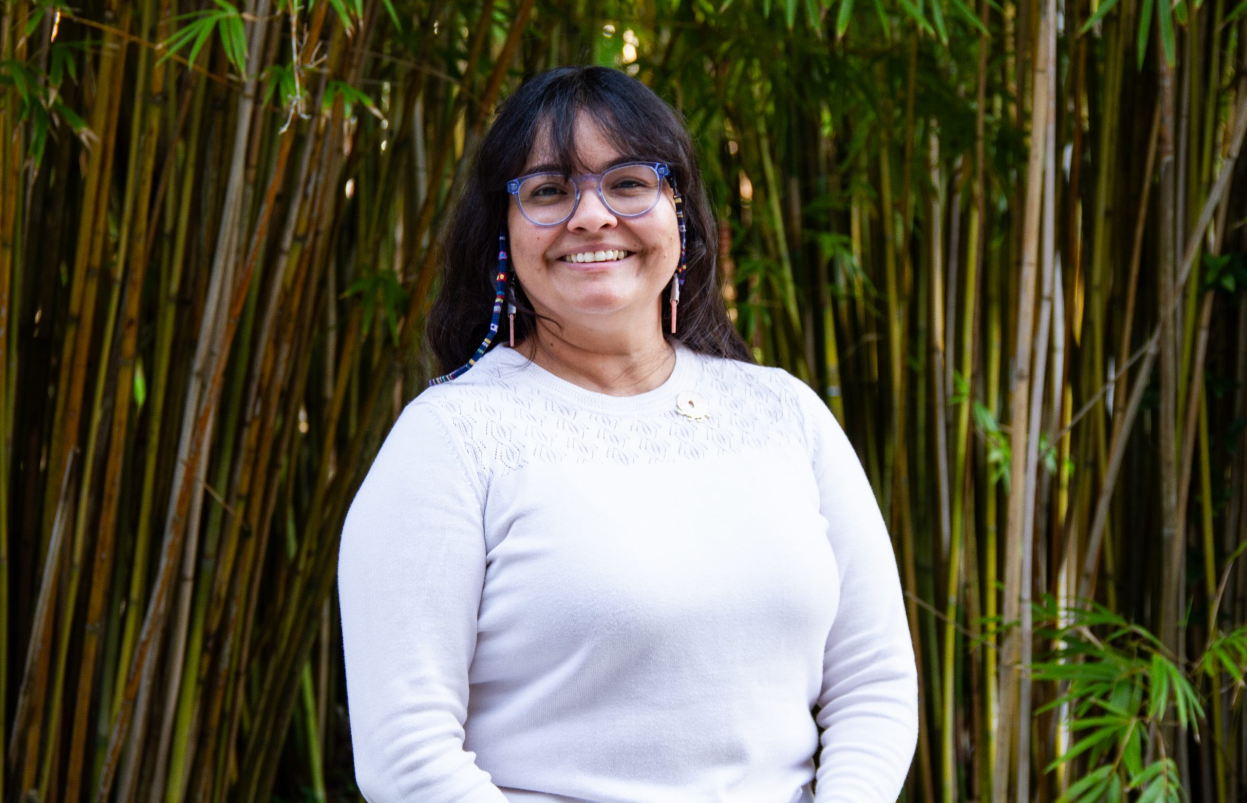 An image of woman with black hair and glasses smiling with bamboo behind her. 