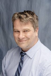 A photo of Bryan. He has  dirty blonde hair and is grinning. He is wearing a blue button up shirt and blue stripey tie. 